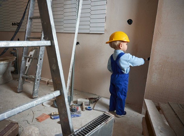 Niño trabajador de la construcción pintando pared con pincel en apartamento en remodelación. Vista posterior del niño con casco de seguridad y mono de trabajo mientras juega en casa. - Foto, imagen