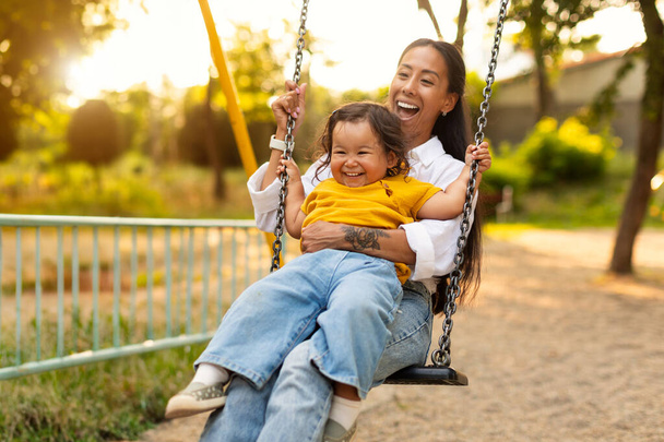 Momenty zabawy. Japońska mama siedzi ze swoją córeczką, swinging Baby i zabawy na placu zabaw na zewnątrz, dzieląc śmiech i radosne więzi w parku na świeżym powietrzu - Zdjęcie, obraz