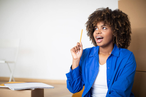 Jeune femme noire excitée ayant une idée tout en faisant une liste de contrôle le jour du déménagement, joyeuse femme afro-américaine levant le crayon et regardant à côté de l'espace de copie, assis parmi les boîtes en carton à la maison - Photo, image