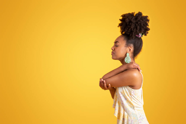 Despaired sad millennial black lady presses hand to elbow, suffer from pain, isolated on orange background, studio, profile. Arthritis, injury and inflammation, health problems, human emotions - Photo, Image
