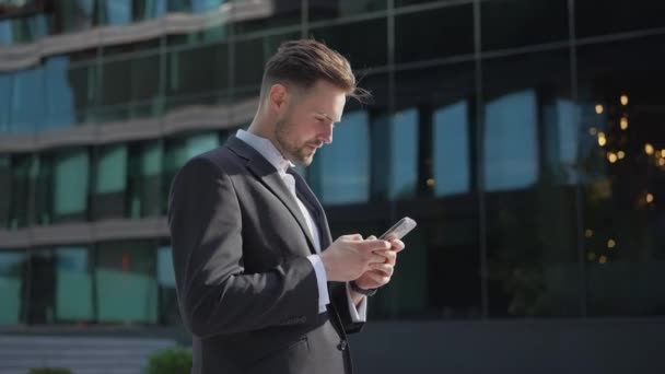 Stylish handsome businessman holding phone reading email stands on street smiling writes message communicates in social network pensive man browses goods in online store using mobile app on smartphone - Footage, Video