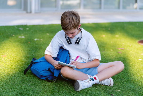 Ragazzo adolescente che fa i compiti a scuola con un quaderno mentre si siede sull'erba all'aperto. Concetto educativo. - Foto, immagini
