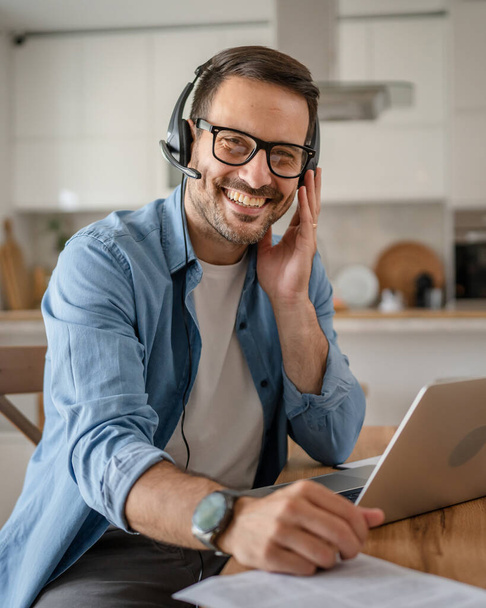 Un homme caucasien travail indépendant masculin de la maison avec des écouteurs sur sa tête sur ordinateur portable répartiteur de soutien à la clientèle sourire heureux confiant - Photo, image