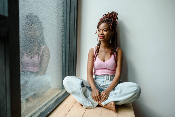 Atractiva mujer africana sonriente con camiseta acanalada y vaqueros sentados con las piernas cruzadas en el alféizar de la ventana y mirando a través de la ventana. Concepto de vida urbana, soñar despierto. Planificación del concepto futuro - Foto, imagen