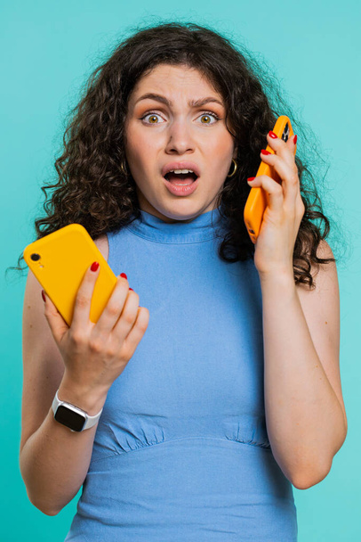 Irritated nervous stressed young woman talking screaming on two mobile phones having conversation conflict quarrel complaint dispute discuss solve problem. Girl on blue studio background. Vertical - Photo, Image