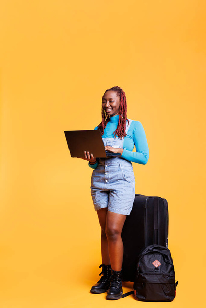 Afroamerykanka pracująca na laptopie, przeglądająca stronę internetową w pomarańczowym studio. Kobieta podróżnik dziewczyna za pomocą komputera do przeglądania strony internetowej online, mając plecak i torby wózek. - Zdjęcie, obraz