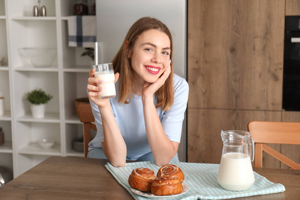 Młoda kobieta ze szklanką mleka i smacznymi bułeczkami w kuchni - Zdjęcie, obraz