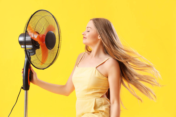 Mujer joven con ventilador eléctrico sobre fondo amarillo - Foto, imagen