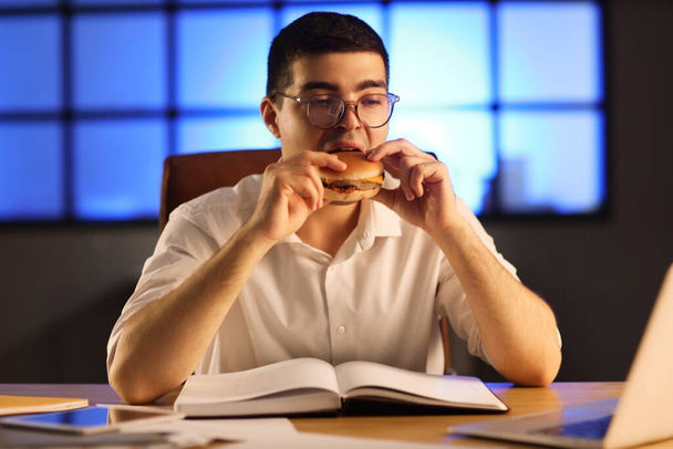 Молодой бизнесмен ест вкусные бургеры в офисе по ночам - Фото, изображение