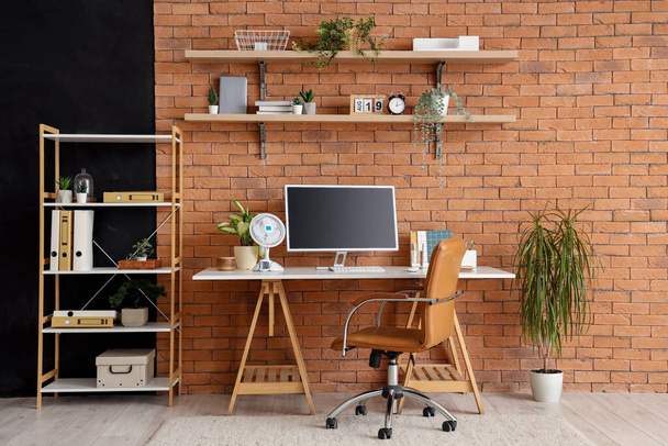 Espace de travail confortable avec ordinateur moderne et ventilateur électrique près du mur de briques brunes - Photo, image