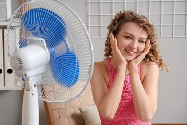 Beautiful happy young woman with electric fan sitting on chair in living room - Photo, Image
