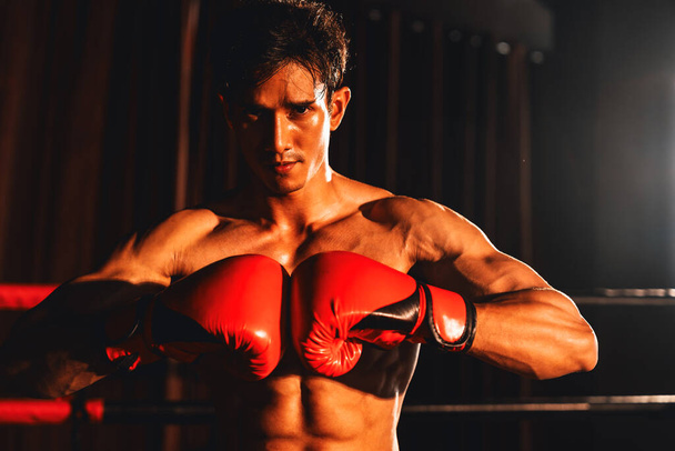 Muay Thai boxer punch fish his hand together in front of camera in ready to fight stance posing at gym with boxing equipment in background. Focused determination eye and prepare for challenge. Impetus - Photo, Image