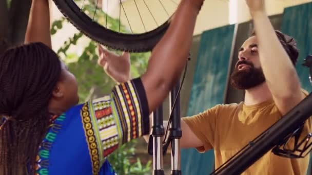 Femme sportive africaine américaine en bonne santé obtient l'aide de l'homme caucasien pour sécuriser une nouvelle roue de vélo dans la cour de la maison. Couple multiethnique amateur de sport refixant pneu de vélo en plein air. - Séquence, vidéo