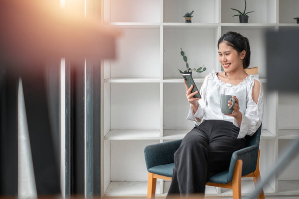 Alegre mujer asiática milenaria usando el teléfono móvil, charlando en la web, trabajando o estudiando en línea, sentado en sillón contra la pared blanca, espacio de copia. - Foto, imagen
