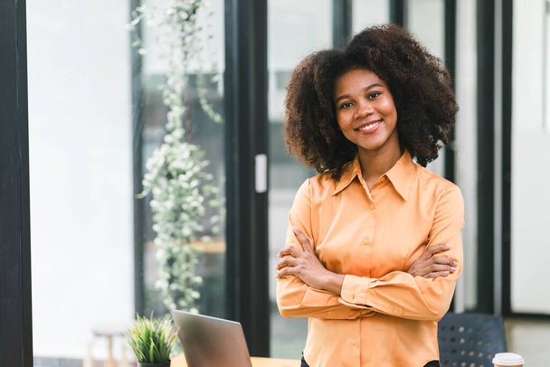 A sikeres és magabiztos fekete üzletasszony portréja. Egy gyönyörű, fiatal afro-amerikai nő mosolyog a kamerába, miközben keresztbe tett kézzel áll egy modern irodában. - Fotó, kép