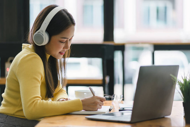 Εστιασμένη Ασιάτισσα επιχειρηματίας φορώντας ακουστικά κρατά σημειώσεις σε ένα σημειωματάριο, ενώ βλέποντας ένα webinar πορεία βίντεο. Σοβαρή φοιτήτρια που ακούει τη διάλεξη για να σπουδάσει online μέσω e-learning - Φωτογραφία, εικόνα