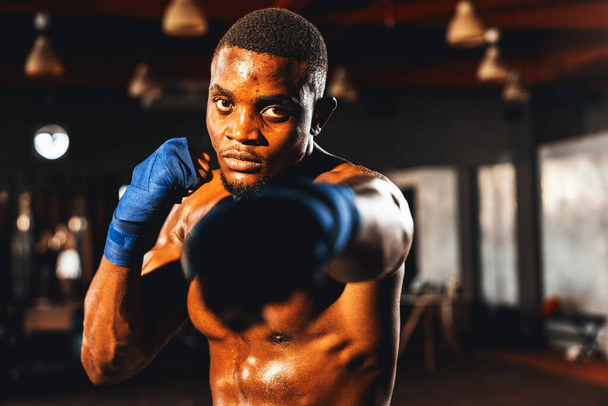 Boxkämpfer in hemdsloser Pose, afroamerikanischer schwarzer Boxer, der seine Faust in aggressiver Haltung schlägt und bereit ist, im Fitness-Studio zu kämpfen, im Hintergrund Trettasche und Boxausrüstung. Impulse - Foto, Bild