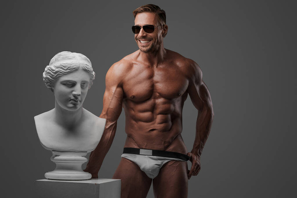Улыбающийся привлекательный мускулистый мужчина-модель в солнечных очках и нижнем белье уверенно стоит рядом с древнегреческим бюстом на сером фоне - Фото, изображение