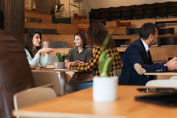 Όμορφη και νεαρή μελαχρινή επιχειρηματίας λέει μια αστεία ιστορία στους φίλους της στην καφετέρια - Φωτογραφία, εικόνα