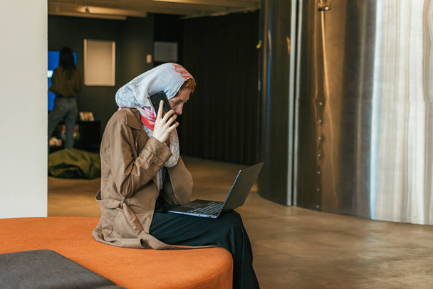 Turuncu koltukta oturan kadın çalışan dizüstü bilgisayarla uğraşırken telefonla konuşuyor. - Fotoğraf, Görsel
