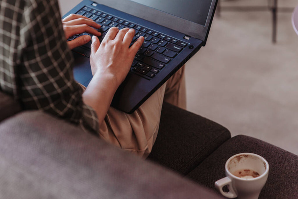 Female employee working on lap top while sitting on a grey sofa with a cup of coffee next to her. Close up photo - Photo, Image
