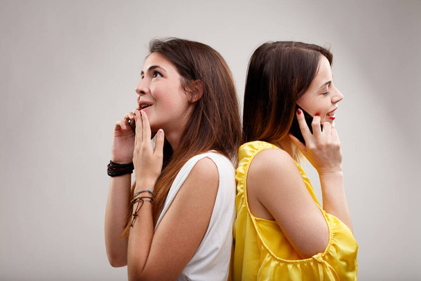 μια νεαρή γυναίκα μοιράζεται πικάντικα κουτσομπολιά με μια άλλη, χλευάζοντας τους άλλους και αναζητώντας έκπληξη για ασυνήθιστες ιστορίες. Γελάνε και χρησιμοποιούν smartphones - Φωτογραφία, εικόνα