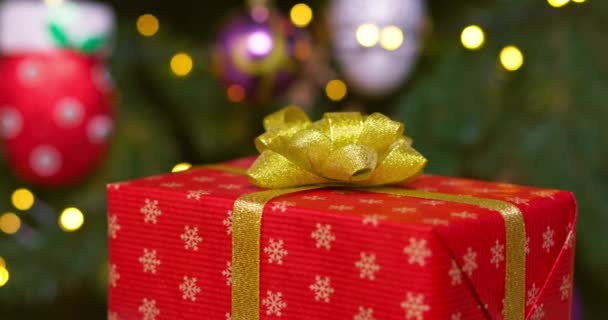 Una caja roja con una cinta dorada y un lazo gira sobre el fondo de un árbol de Navidad con luces de guirnalda intermitentes. Imágenes de alta calidad 4k - Metraje, vídeo