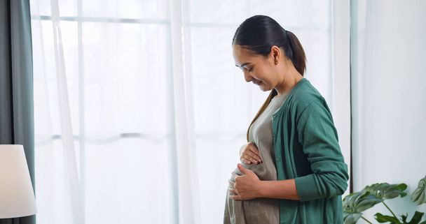 Onnellinen aasialainen raskaana oleva nainen koskettaa rakastavasti syntymätöntä lastaan seistessään talon ikkunan vieressä.. - Valokuva, kuva