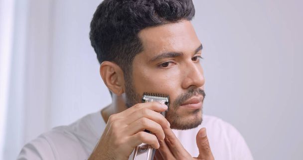 Un uomo sta usando rasoio elettrico per tagliare i baffi e la barba mentre in piedi davanti allo specchio in bagno a casa. - Foto, immagini