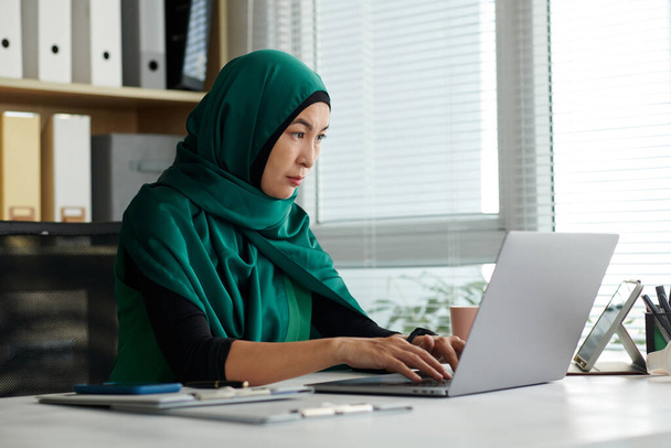 Mujer musulmana seria en hijab trabajando en la computadora portátil en la oficina - Foto, imagen