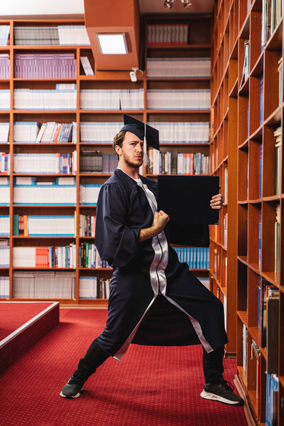 Junger und aufgeregter Absolvent mit Mütze und Kleid feiert seinen Bachelor-Abschluss in einer Bibliothek - Foto, Bild