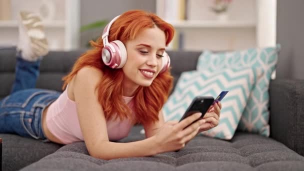 Fiatal vörös hajú nő vásárol okostelefonnal és hitelkártyával zenét hallgat otthon - Felvétel, videó