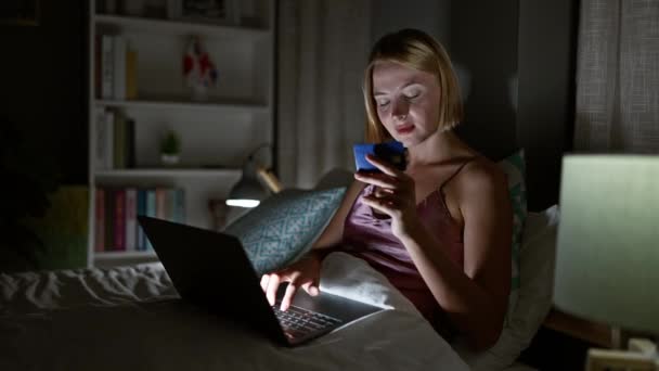 Νεαρή ξανθιά γυναίκα ψώνια με φορητό υπολογιστή και πιστωτική κάρτα κάθεται στο κρεβάτι στο υπνοδωμάτιο - Πλάνα, βίντεο