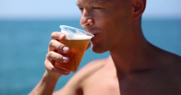 Mies juo olutta rannalla muovikupista. Ranta puolue ja kylmä olut matkailijoille - Materiaali, video
