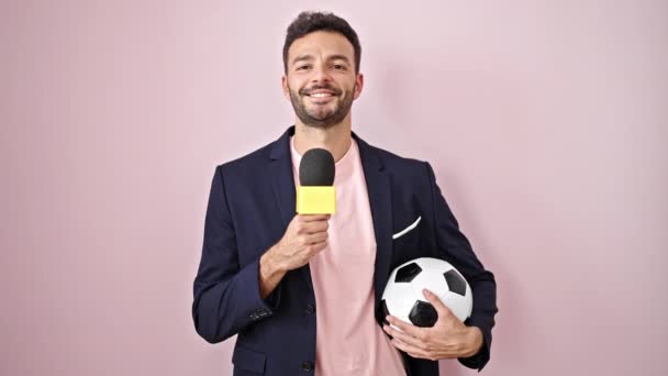 Fiatal spanyol férfi foci riporter kezében mikrofon és labda mosolyog izolált rózsaszín háttér - Felvétel, videó