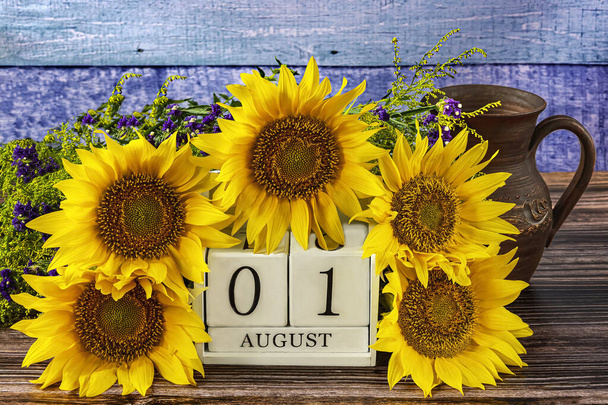 Перший серпень на білому дерев'яному календарі і красиві жовті квіти соняшнику навколо, глечик на синьому дерев'яному фоні. - Фото, зображення