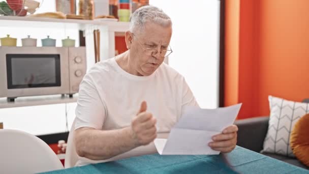 Μέση ηλικία γκριζομάλλης άνθρωπος ανάγνωση έγγραφο κάθεται στο τραπέζι με την έκφραση νικητής στην τραπεζαρία - Πλάνα, βίντεο