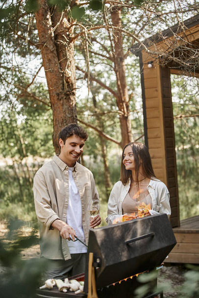 Coppia allegra con vino che cucina sulla griglia durante il picnic vicino alla casa estiva sullo sfondo - Foto, immagini