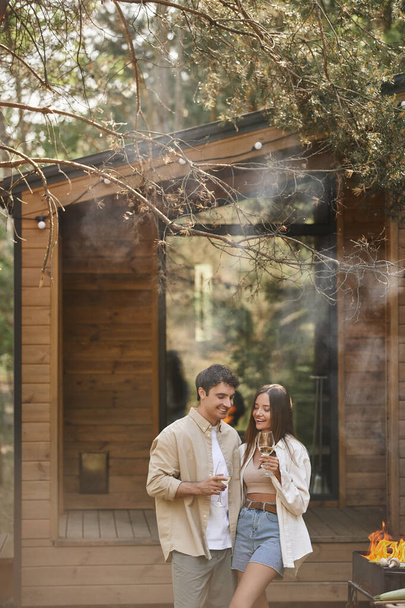 Cheerful and stylish couple holding wine and standing near grill with smoke and vacation house - Foto, Bild