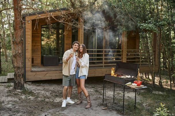 Positive romantic couple holding wine and looking at camera near grill and vacation house - Photo, Image