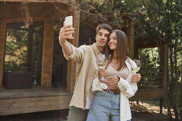 Κομψός άνθρωπος λαμβάνοντας selfie στο smartphone με τη φίλη κρατώντας κρασί κοντά στο σπίτι το καλοκαίρι στο παρασκήνιο - Φωτογραφία, εικόνα