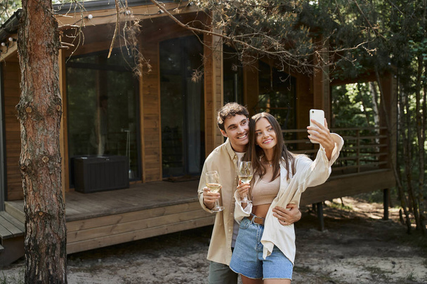 Smiling man hugging girlfriend and taking selfie with girlfriend holding wine near vacation house - Foto, afbeelding