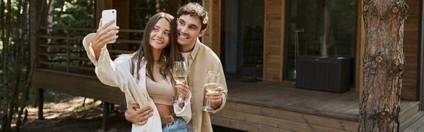 Hymyilevä romanttinen pari viiniä ottaen selfie älypuhelimeen lähellä hämärtynyt kesämökki, banneri - Valokuva, kuva