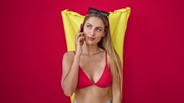 Junge blonde Touristin im Bikini unterhält sich auf Smartphone vor isoliertem roten Hintergrund - Filmmaterial, Video
