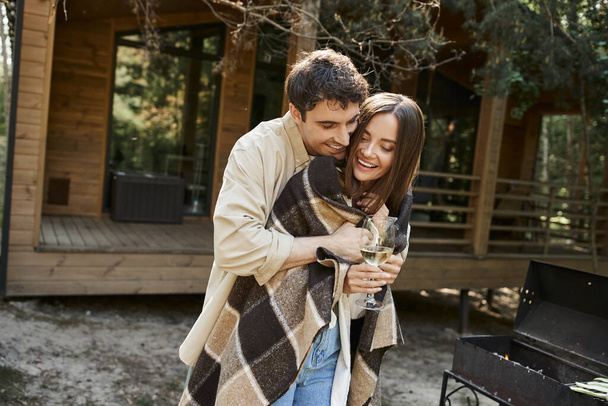 Fröhlicher Mann umarmt Freundin in Decke mit Wein neben Grill und Ferienhaus im Hintergrund - Foto, Bild