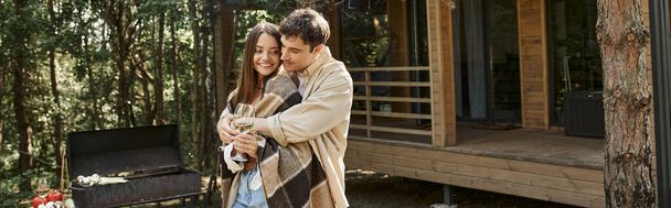 Positive man hugging girlfriend in blanket with wine near grill and summer house, banner - Photo, Image