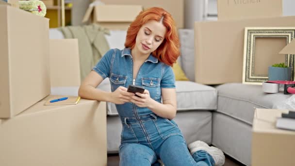 Młoda ruda kobieta za pomocą smartfona siedzi na podłodze w nowym domu - Materiał filmowy, wideo