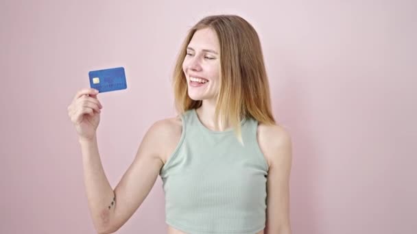 fiatal szőke nő mosolygós magabiztos gazdaság hitelkártya győztes gesztus felett elszigetelt rózsaszín háttér - Felvétel, videó