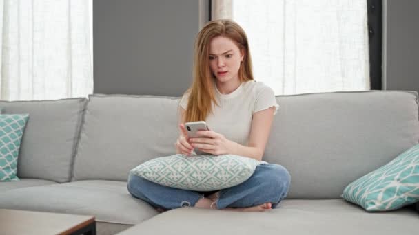 Νεαρή ξανθιά γυναίκα ρίχνουν smartphone κάθεται στον καναπέ τόνισε στο σπίτι - Πλάνα, βίντεο