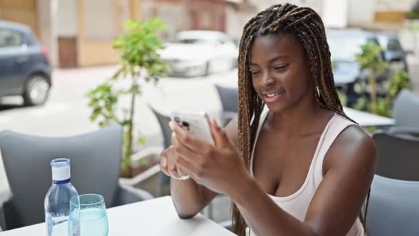 African american woman using smartphone sitting on table with winner gesture at coffee shop terrace - Footage, Video